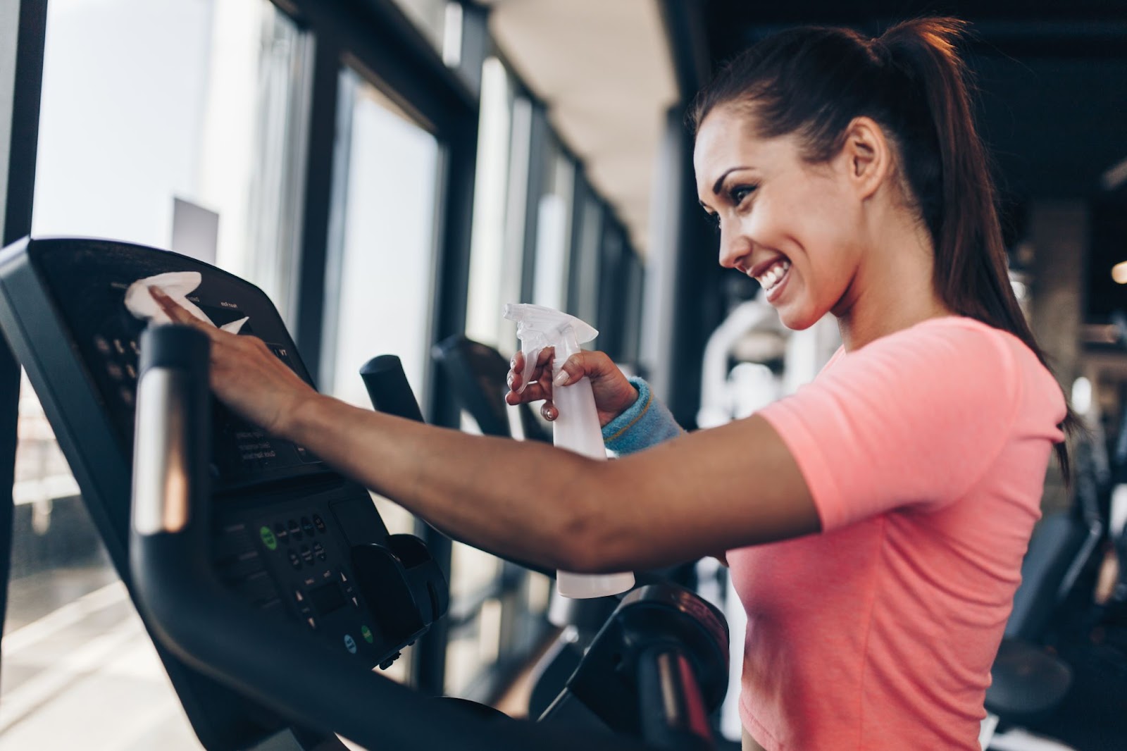 Smiling young woman disinfects and cleans gym equipment in fitness facility.